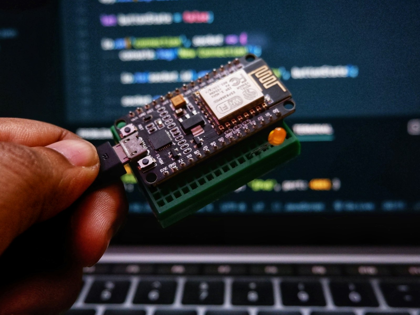 ESP8266 microcontroller board held up in front of a laptop running code